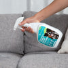 A hand spraying on a grey couch using the Potty Buddy biological stain and odor cleaning spray